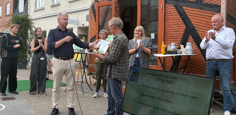 Torben Wallner overrækker Grøn Skanderborg Prisen til Henrik Borgtoft foran Klimakaravanen. Skærm med afstemningsmulighed ses i forgrunden og de to andre nominerede står bagved og klapper af Henrik. 